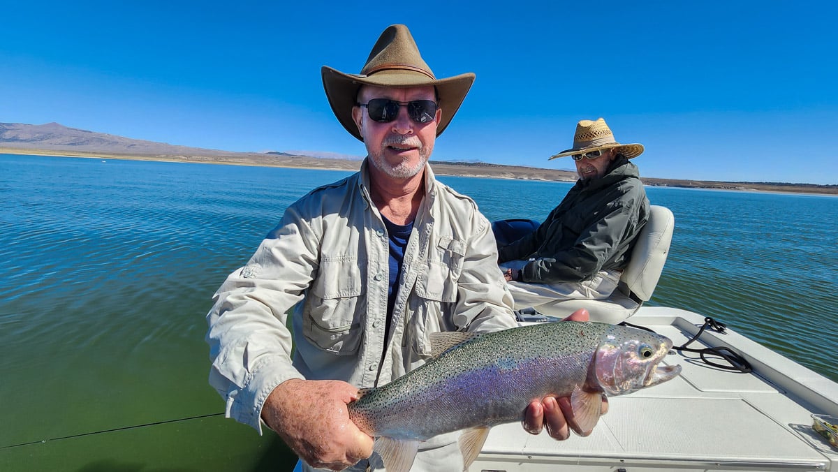 A smiling fly fisherman on a lake in a boat holding a large rainbow trout.