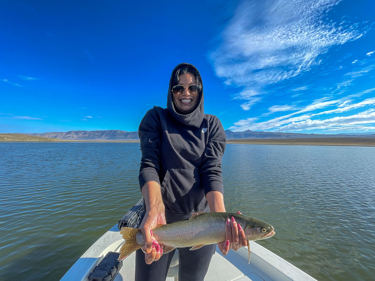 A smiling female fly fisherman on a lake in a boat holding a large cutthroat trout.