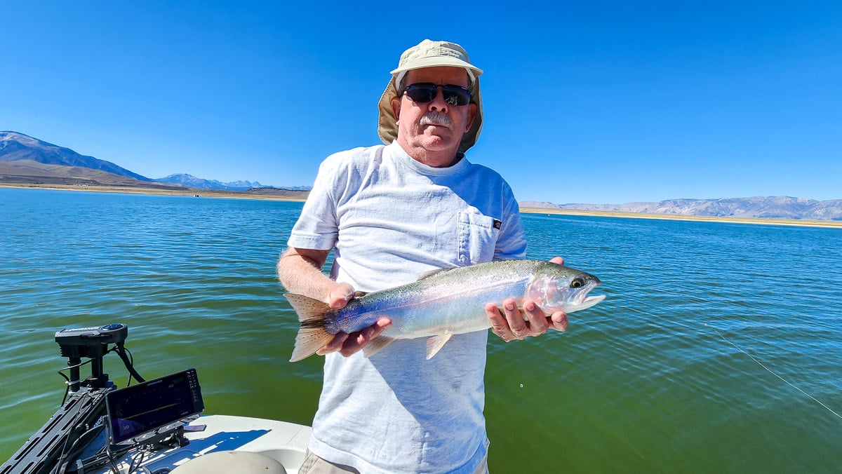 A smiling fly fisherman on a lake in a boat holding a large rainbow trout.