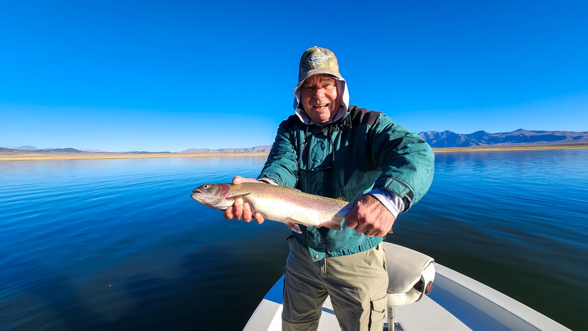 A smiling fly fisherman on a lake in a boat holding a large cutthroat trout.