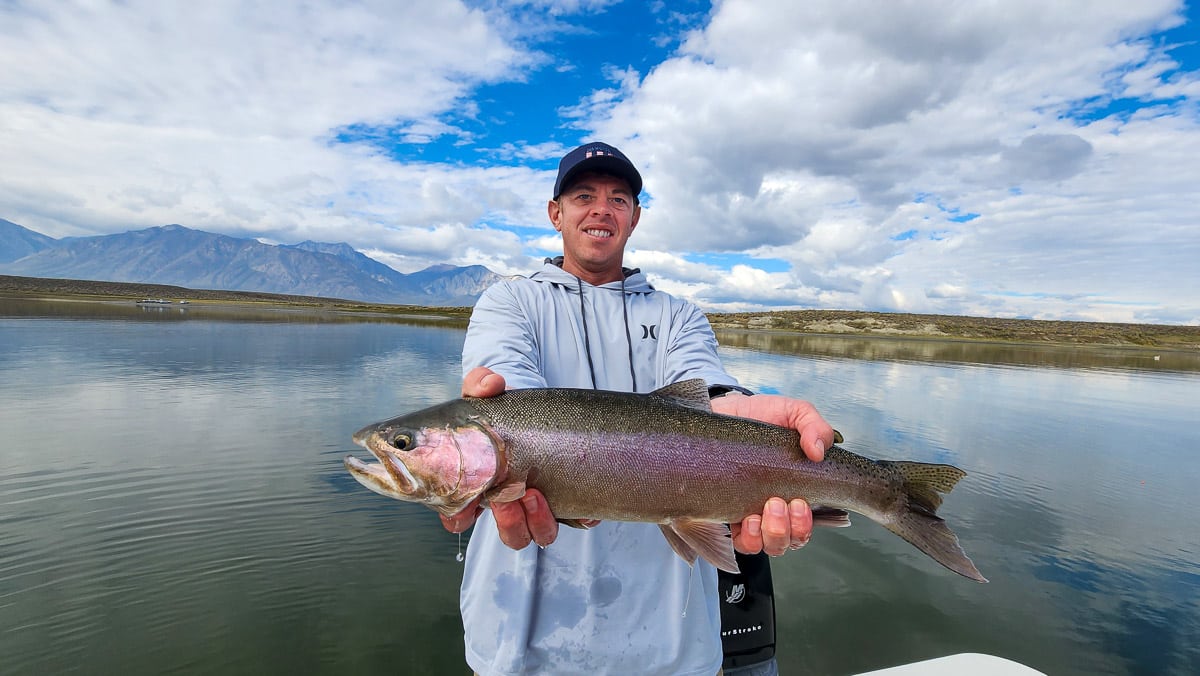 A smiling fly fisherman on a lake in a boat holding a large rainbow trout.