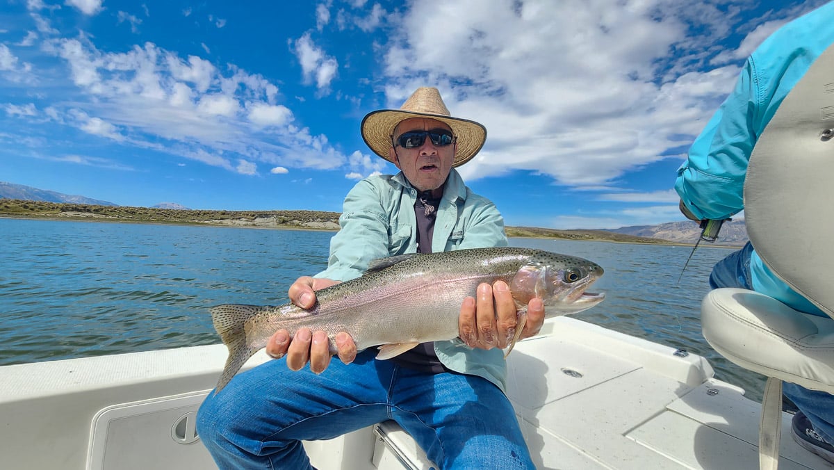 A smiling fly fisherman on a lake in a boat holding a large rainbow trout.