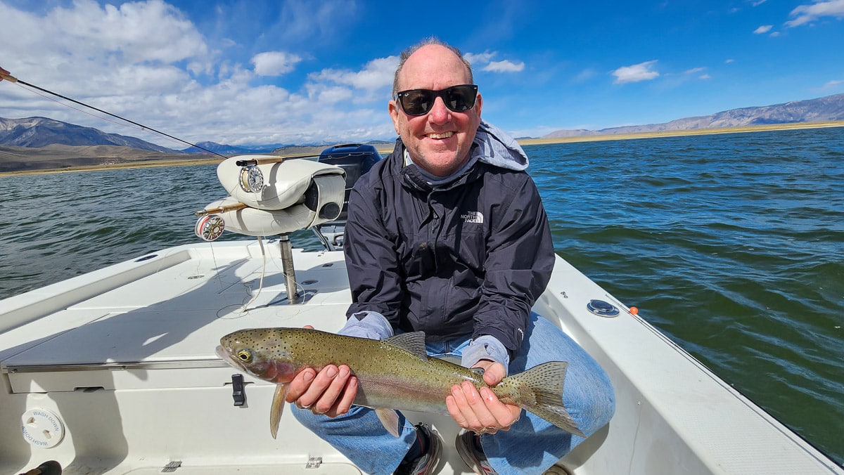 A smiling fly fisherman on a lake in a boat holding a large rainbow trout.