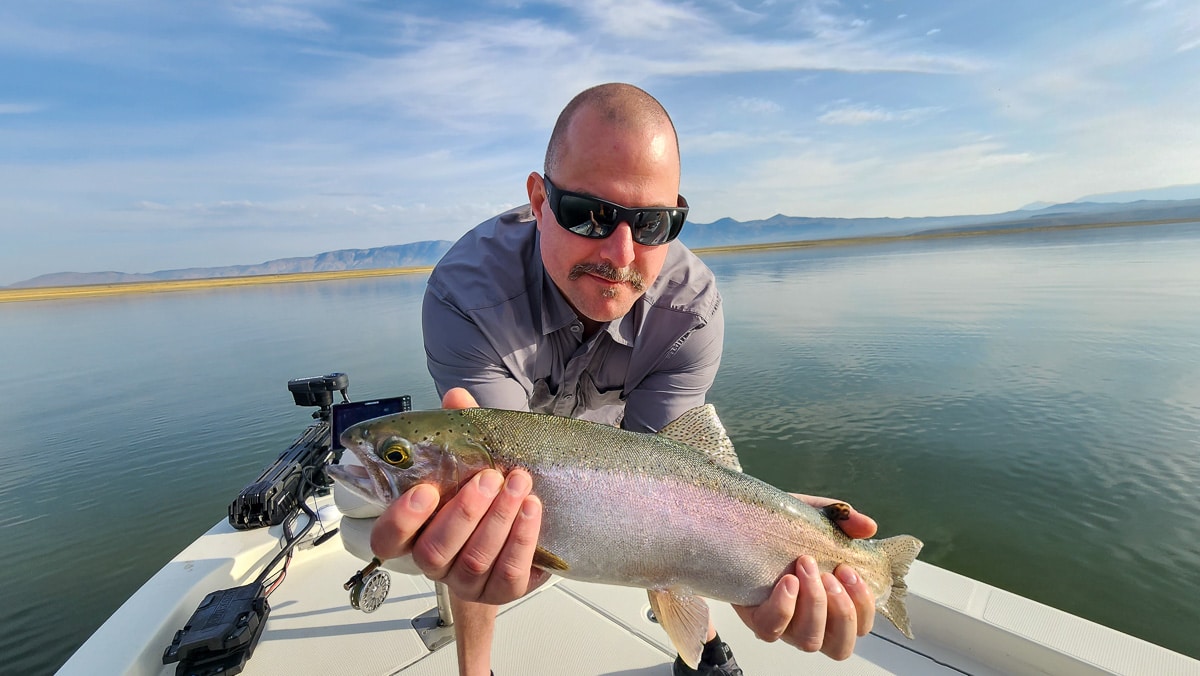 A smiling fly fisherman on a lake in a boat holding a large rainbow trout.