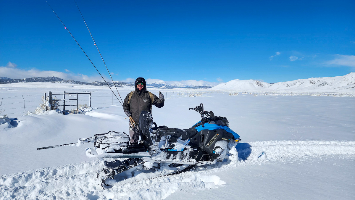 A fisherman standing in the snow next to a blue and black snowmobile which has left its tracks in fresh snow.