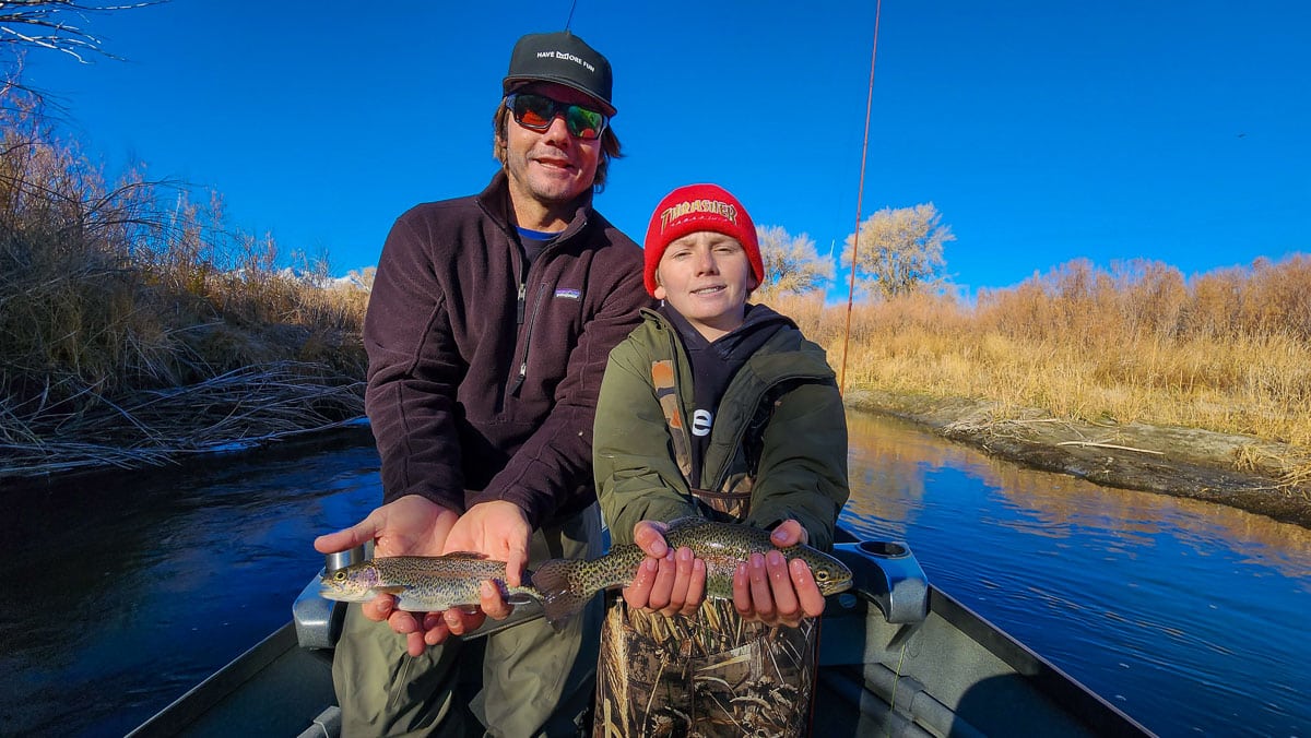 A man and a young boy holding a pair trout in a boat on a river.