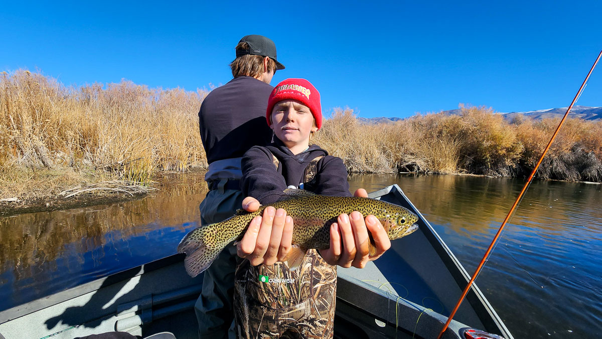 A young boy holding a rainbow trout in a boat on a river with a man behind him fishing.