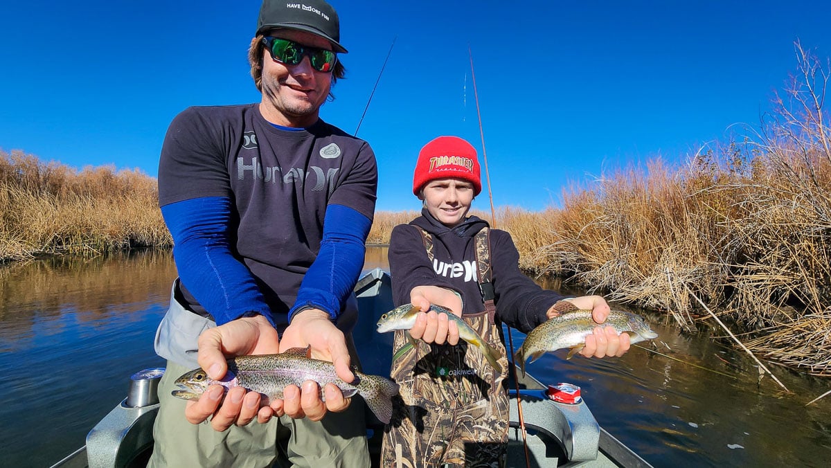 A man and a young boy holding three trout in a boat on a river.