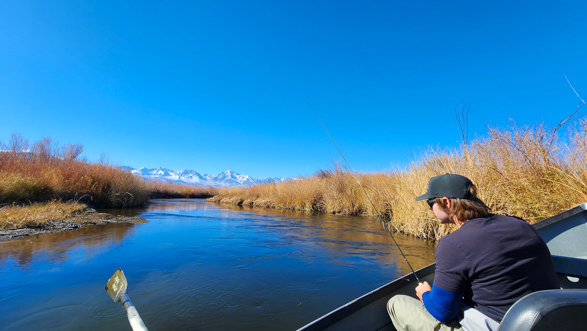 A fisherman in a boat on a river holding a fly fishing rod with snow capped mountains in the background.
