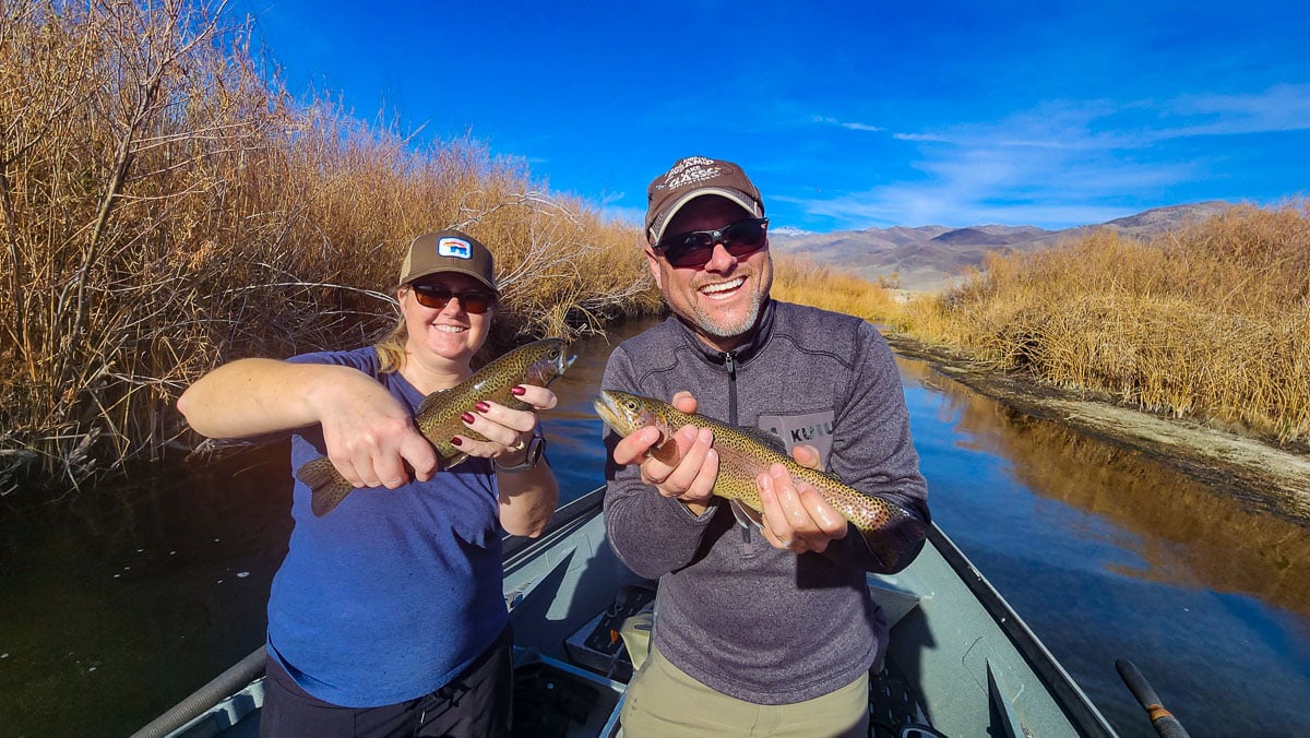 A man and a woman in a boat on a river holding a pair of rainbow trout.