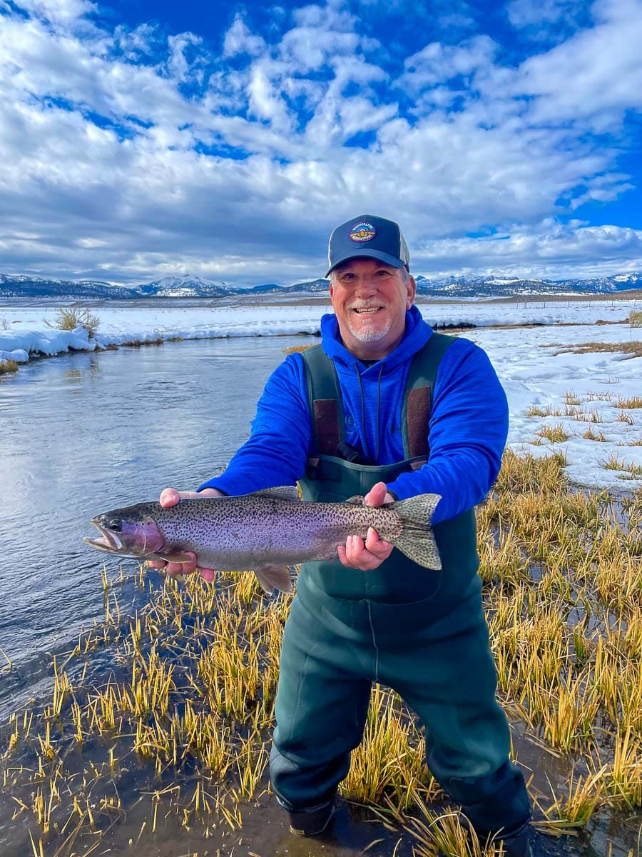 A smiling fisherman holding a massive rainbow trout next to a river in the snow.