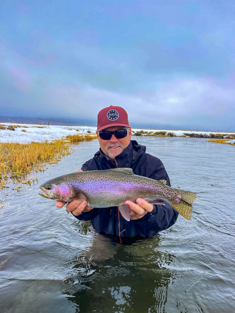 A smiling fisherman holding a massive rainbow trout next to a river in the snow.