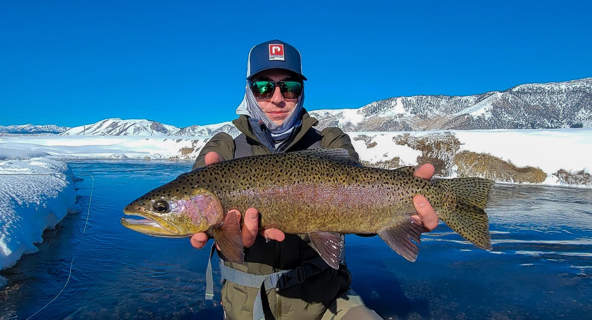 A smiling fisherman holding a massive rainbow trout next to a river in the snow.