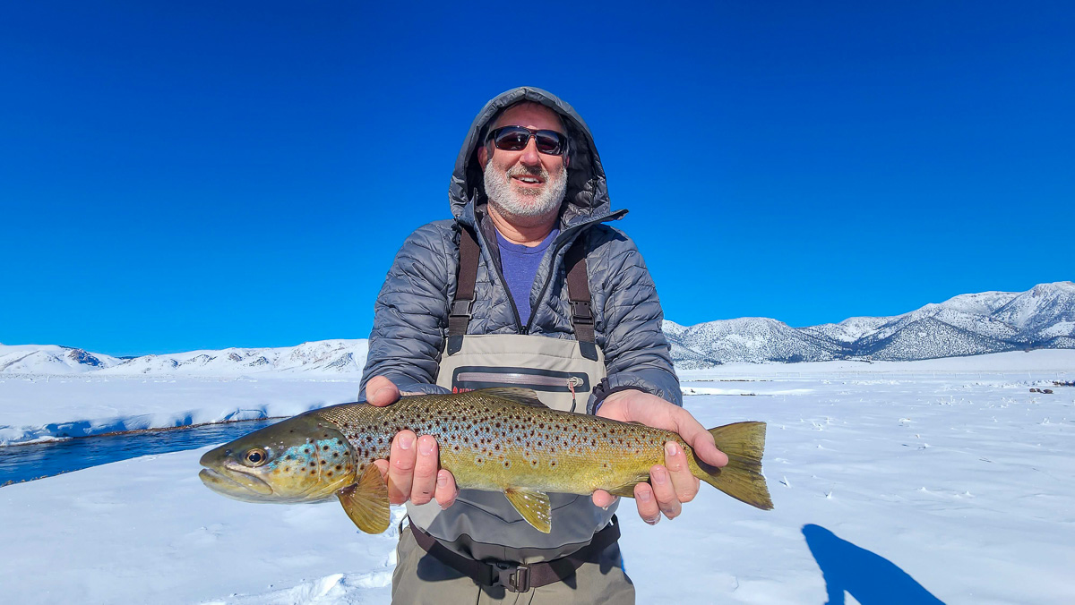 A smiling fisherman holding a massive brown trout next to a river in the snow.