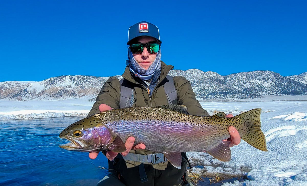 A smiling fisherman holding a massive rainbow trout next to a river in the snow.