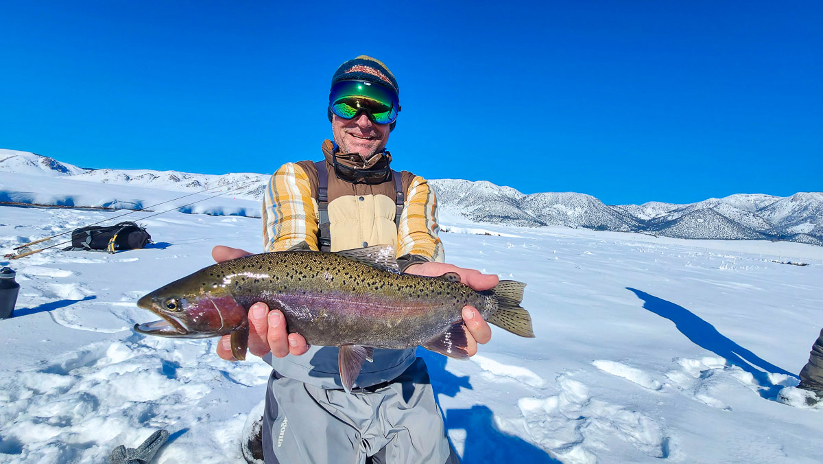 A smiling fisherman holding a massive rainbow trout next to a river in the snow.