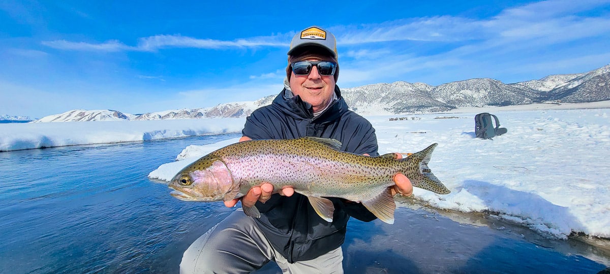 A smiling fisherman holding a massive rainbow trout next to a river in the snow.