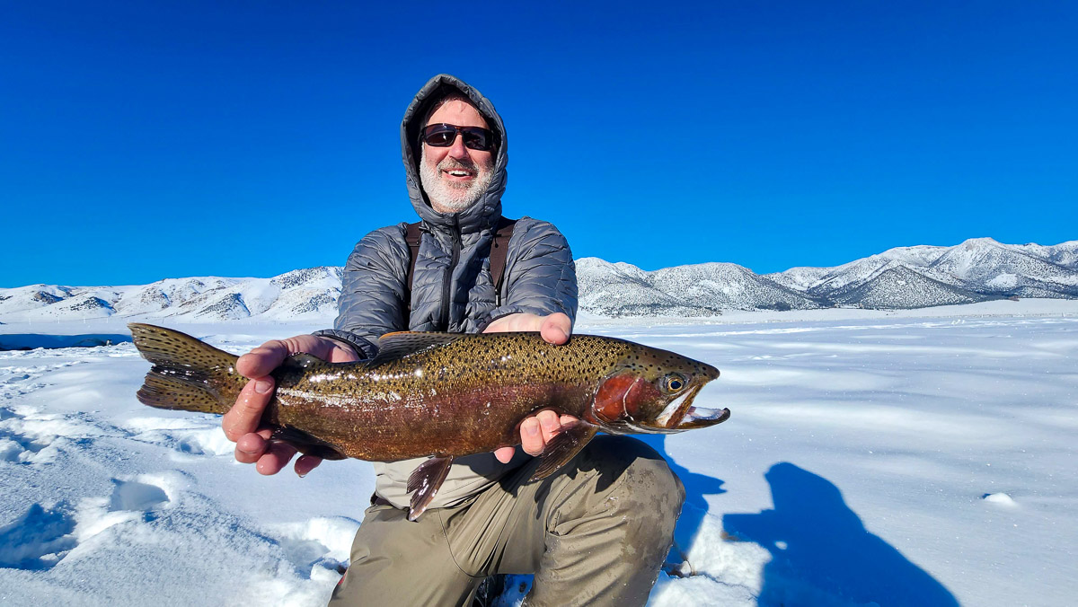 A smiling fisherman holding a massive rainbow trout next to a river in the snow.