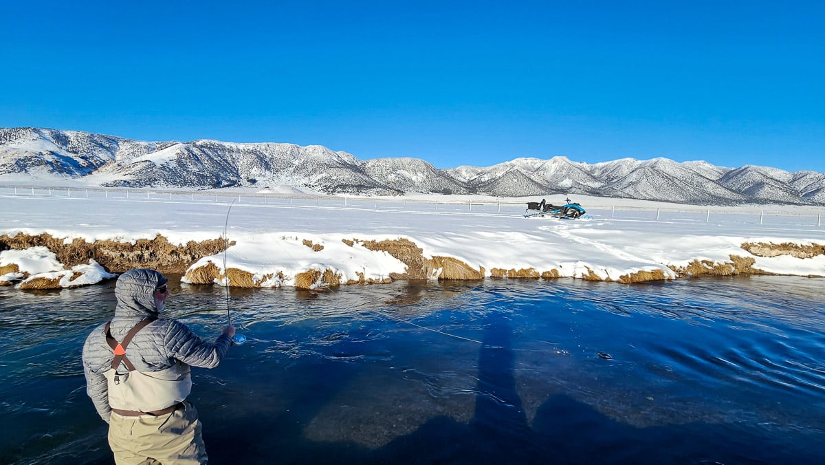 A man in cold weather clothing fly fishing on a river in the winter with snow on the banks.