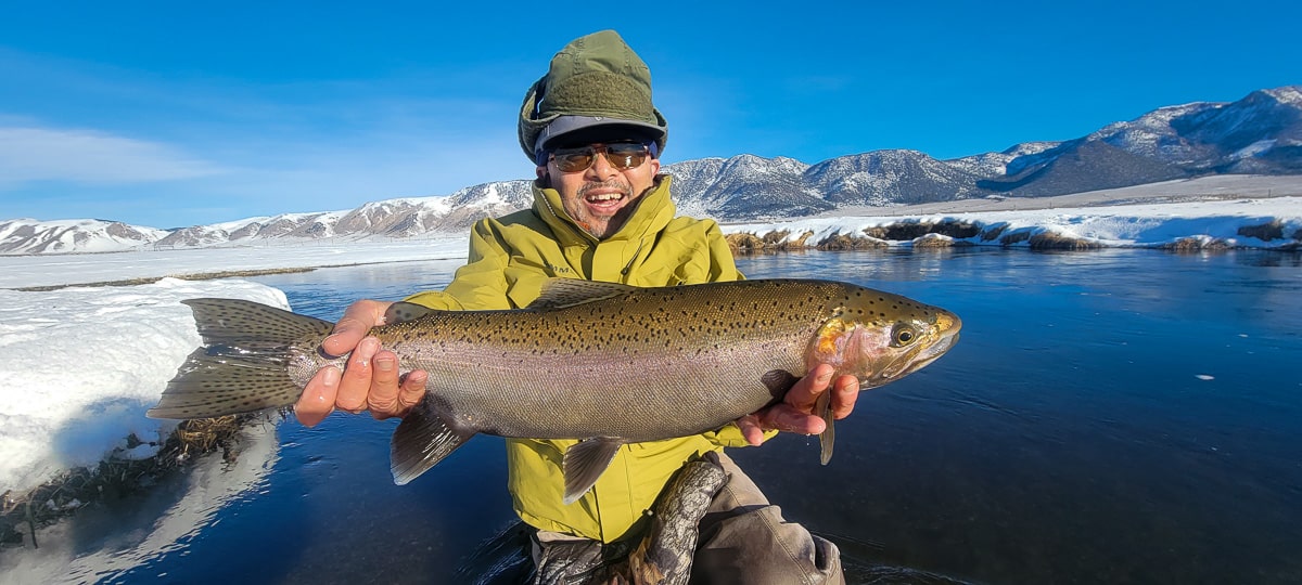 A smiling fisherman holding a massive rainbow trout next to a river in the snow.