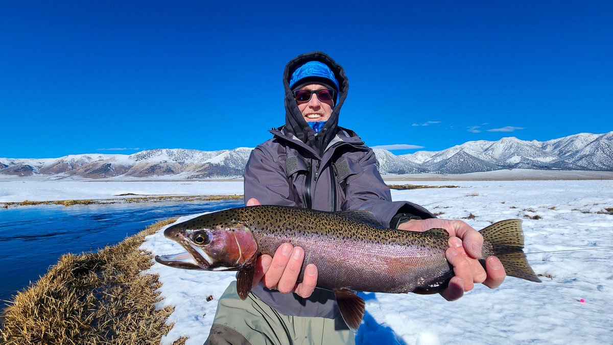 A smiling fisherman holding a massive rainbow trout next to a river in the snow..