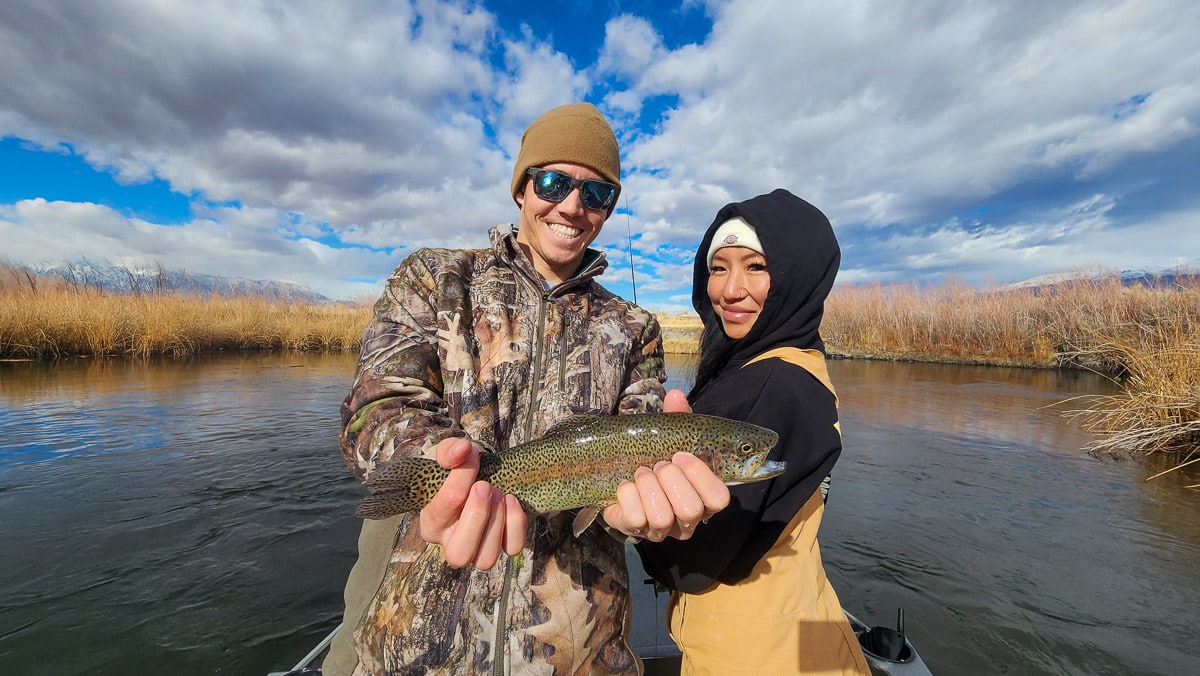 A smiling angler in wearing a brown beanie and a camouflage jacket holding a rainbow trout in a river with a woman wearing a black hooded sweatshirt.