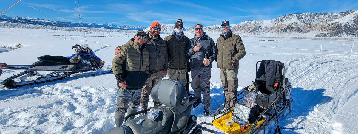 Five men wearing cold weather clothing standing next to snowmobiles in the snow in the mountains.