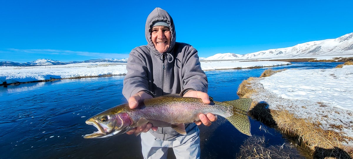 A smiling fisherman holding a massive rainbow trout next to a river in the snow.