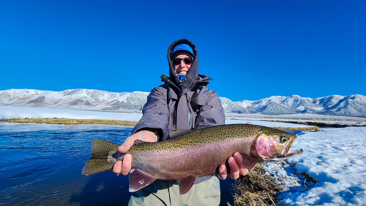 A smiling fisherman holding a massive rainbow trout next to a river in the snow.