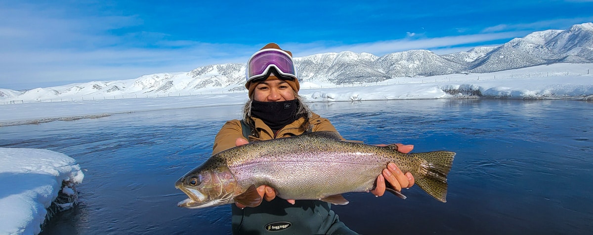 A smiling fisherwoman holding a massive rainbow trout next to a river in the snow in the eastern sierra fishing zone.