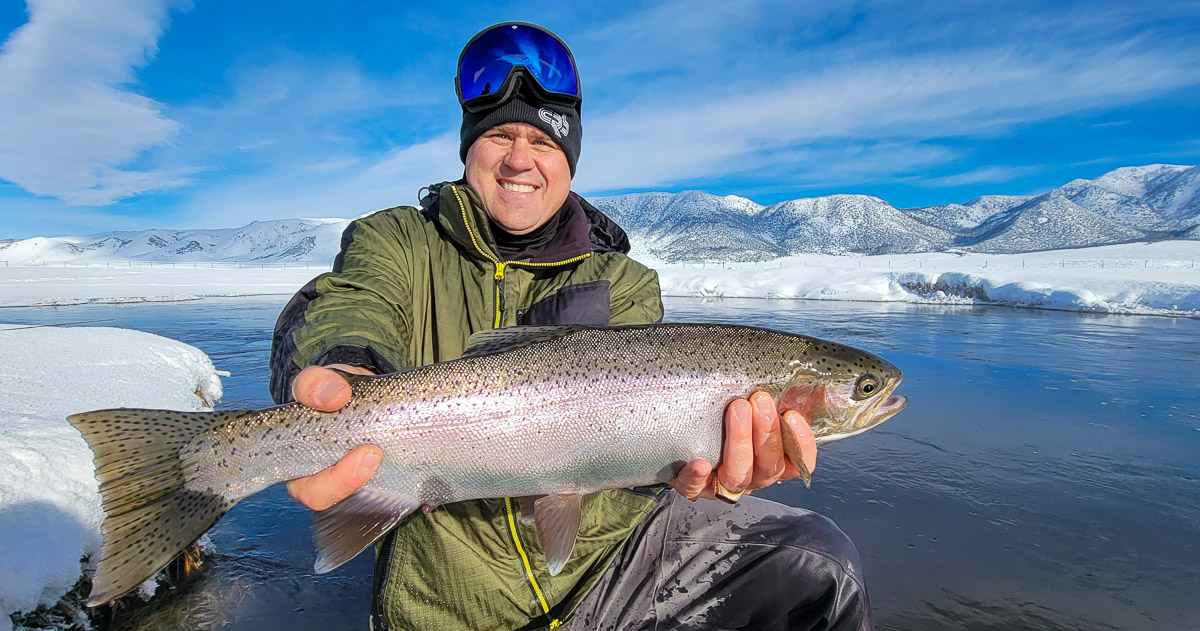 A smiling fisherman holding a rainbow trout in a river in the eastern sierra fishing zone.