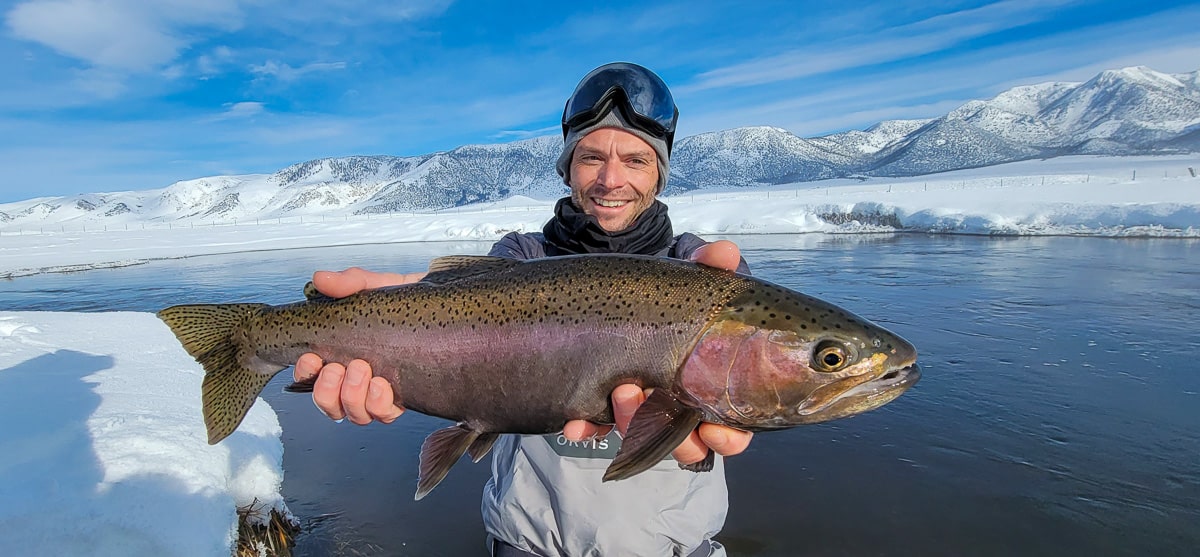 A smiling fisherman holding a massive rainbow trout next to a river in the snow in the eastern sierra fishing zone.