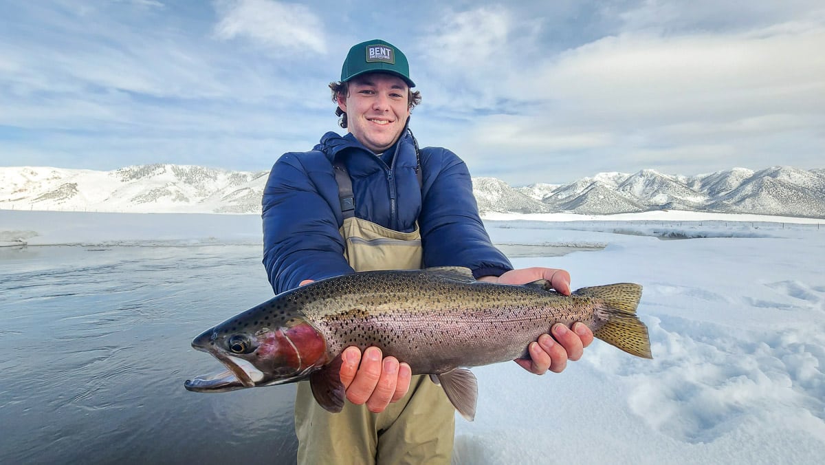 A smiling fisherman holding a massive rainbow trout next to a river in the snow.