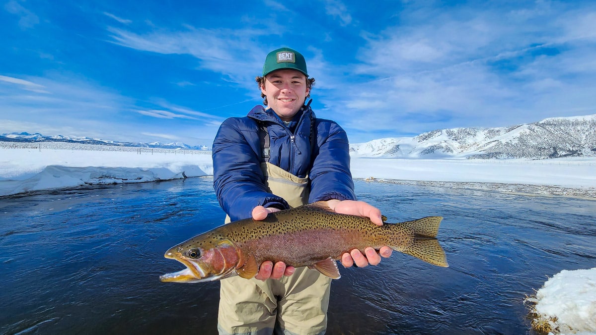 A smiling fisherman holding a massive rainbow trout next to a river in the snow in the eastern sierra fishing zone.