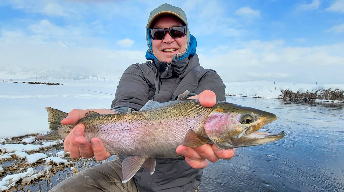 A smiling fisherman holding a massive rainbow trout next to a river in the snow in the eastern sierra fishing zone.