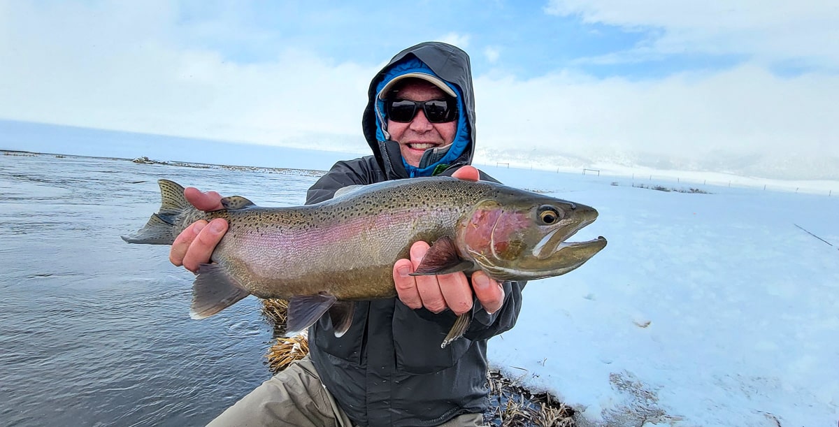 A smiling fisherman holding a massive rainbow trout next to a river in the snow in the eastern sierra fishing zone.