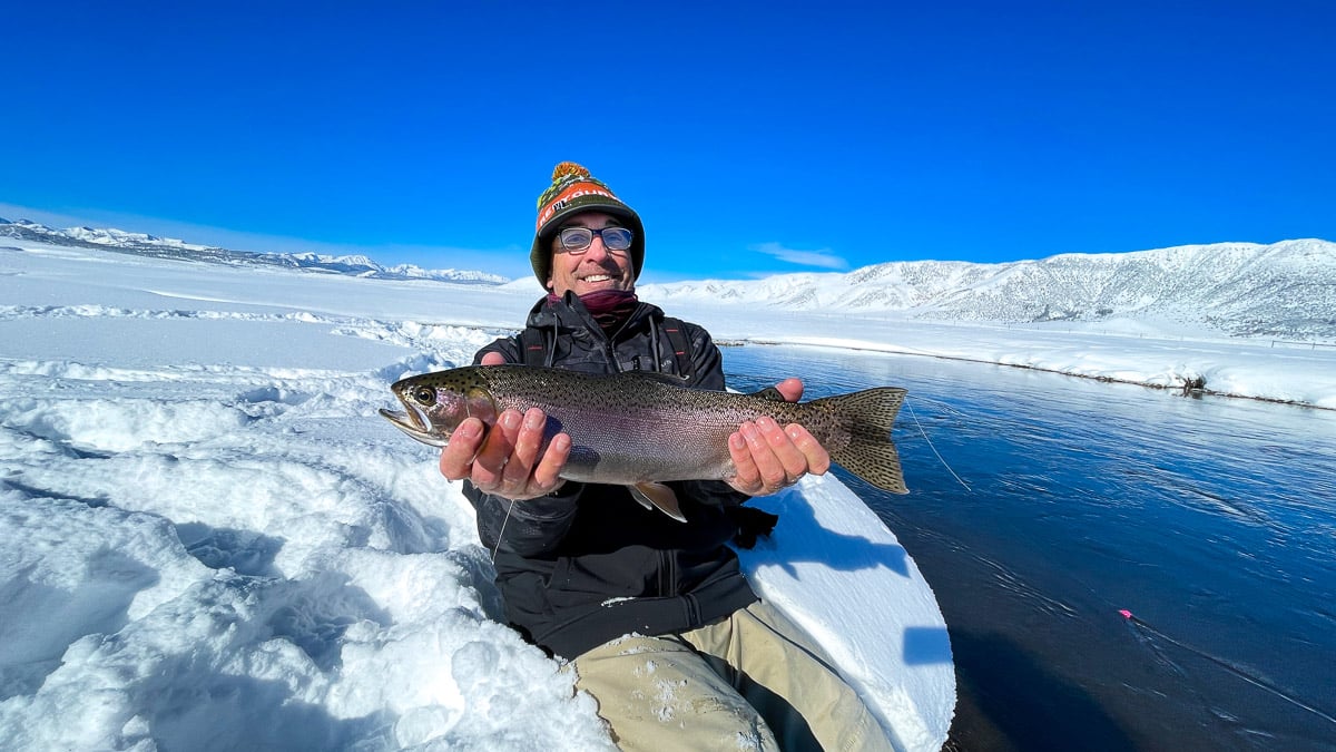 A smiling fisherman holding a massive rainbow trout next to a river in the snow in the eastern sierra fishing zone.