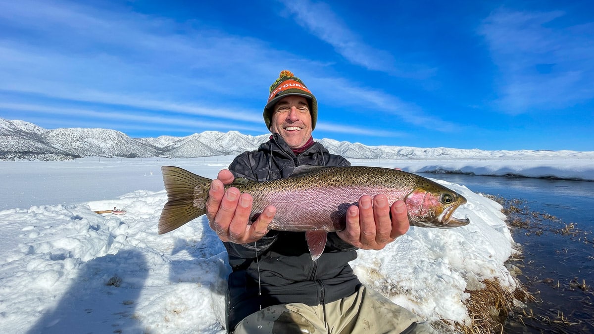 A smiling fisherman holding a massive rainbow trout next to a river in the snow in the eastern sierra fishing zone.