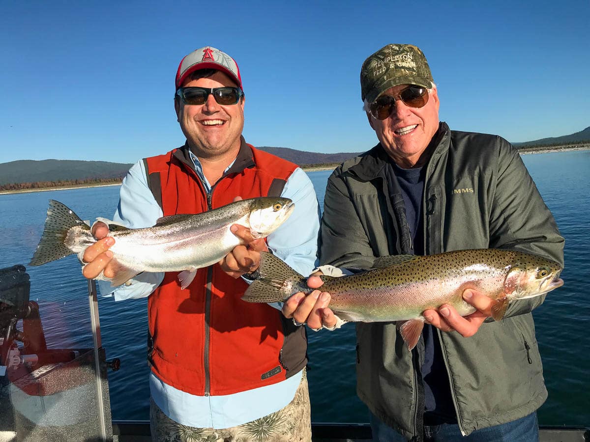 Two men with baseball caps holding very large rainbow trout in a boat on Eagle Lake.