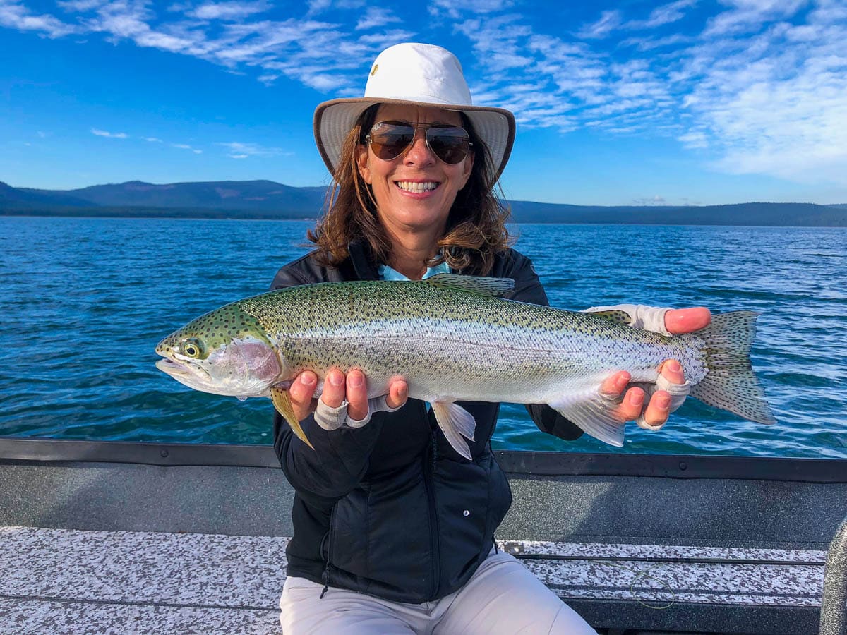 A woman with a white wide brimmed hat and brown lens sunglasses holding a large rainbow trout in a boat on Eagle Lake.