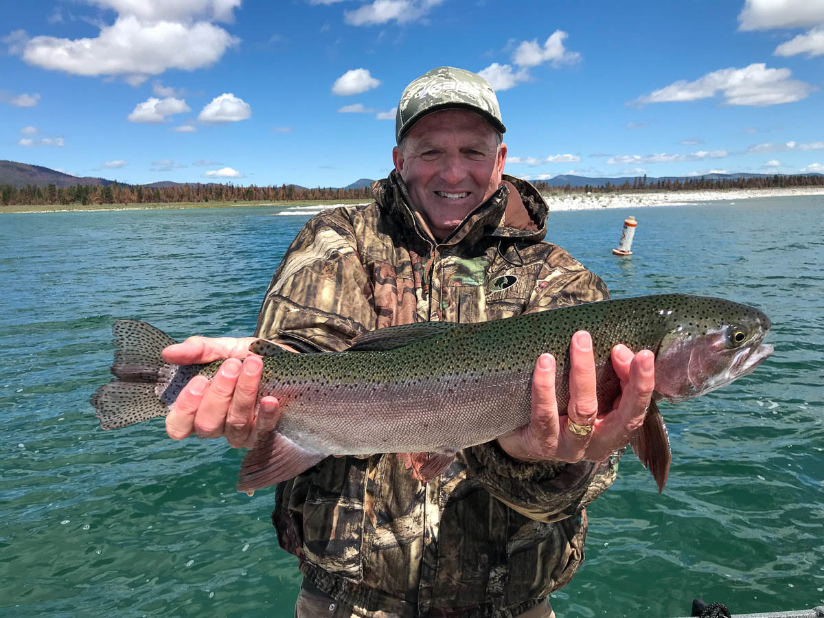 A man with a camouflage baseball hat wearing a camouflage jacket holding a large rainbow trout in a boat on Eagle Lake.