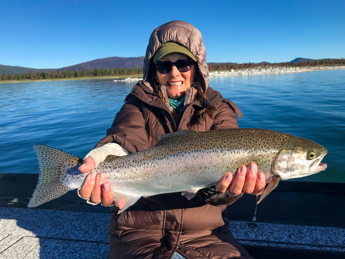A woman with a green baseball cap and a down feathered hood holding a giant rainbow trout in a boat on Eagle Lake.