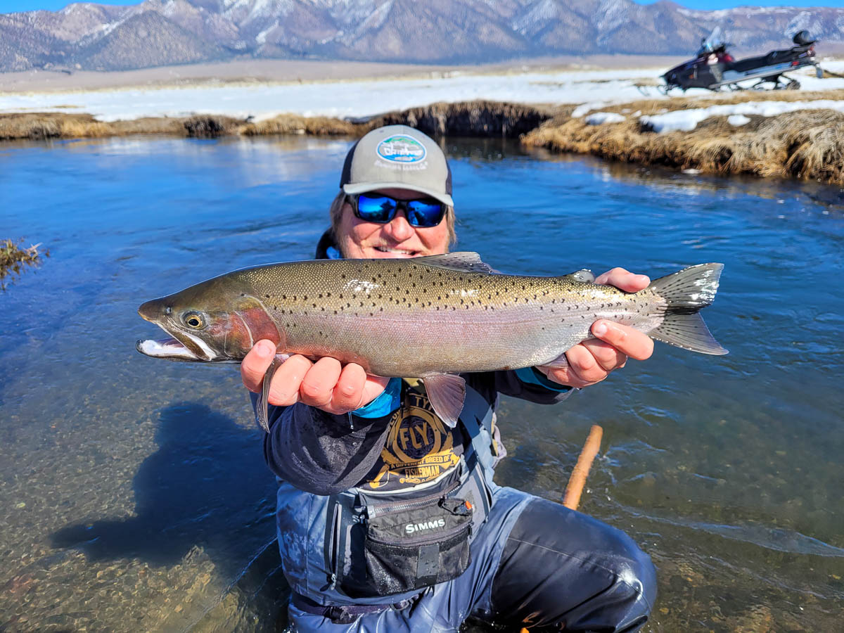 A smiling fisherman holding a massive rainbow trout next to a river in the snow on the Upper Owens River.