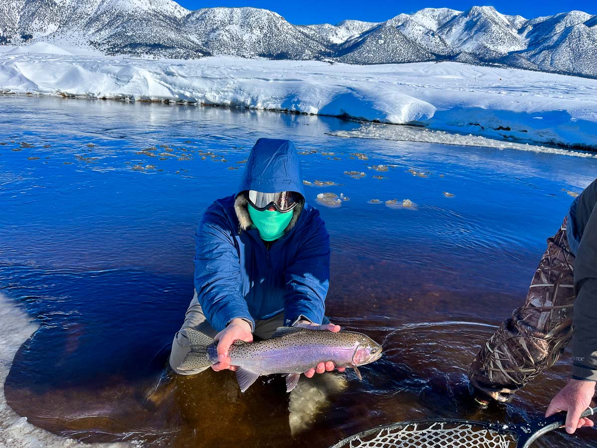 A smiling fisherwoman holding a massive rainbow trout next to the Upper Owens River in the snow in the eastern sierra fishing zone.