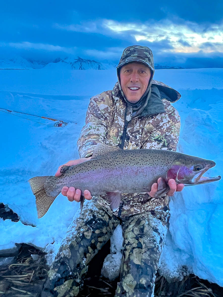 A smiling fisherman holding a massive rainbow trout next to a river in the snow in the eastern sierra fishing zone.