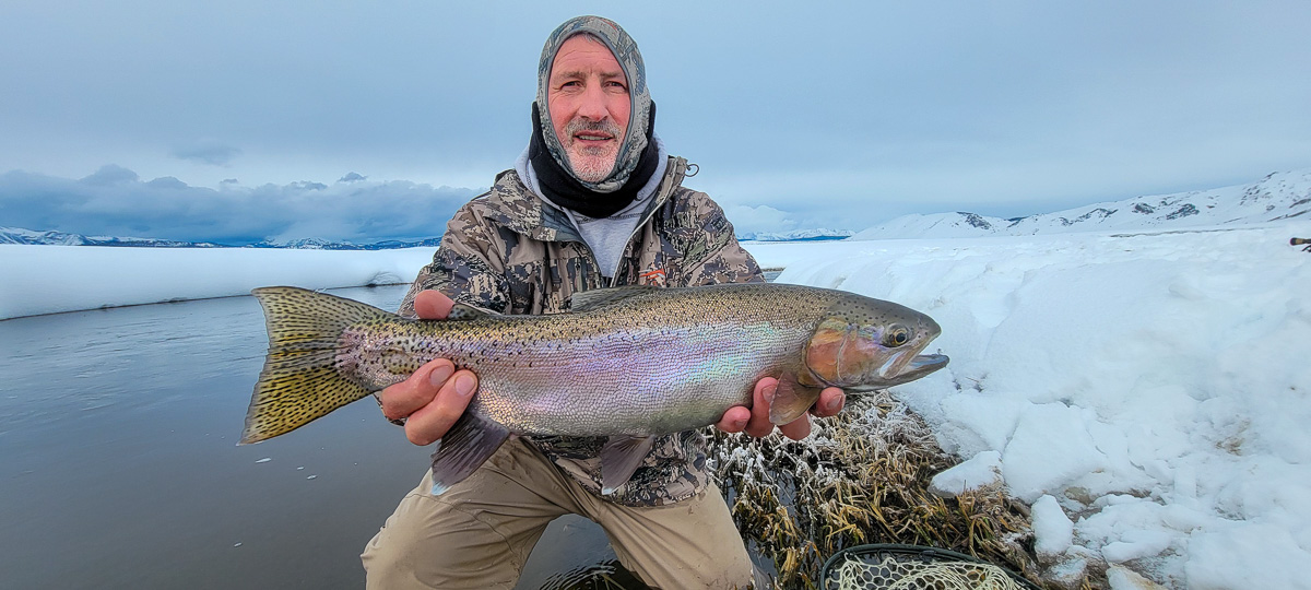 A fisherman holding a massive rainbow trout in the snow by a river.