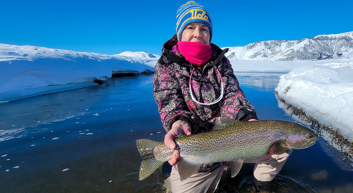 A smiling lady fisherman holding a massive rainbow trout next to a river in the snow on the Upper Owens River.