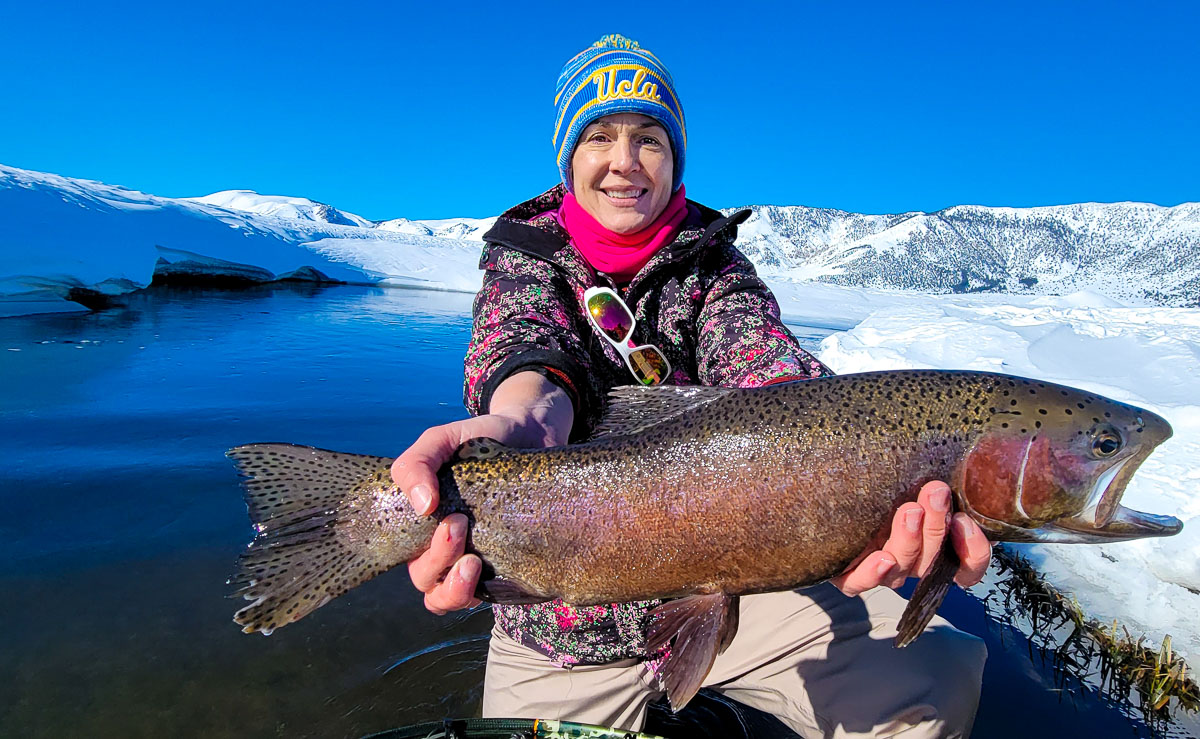 A smiling fisherwoman holding a massive rainbow trout next to the Upper Owens River in the snow in the eastern sierra fishing zone.