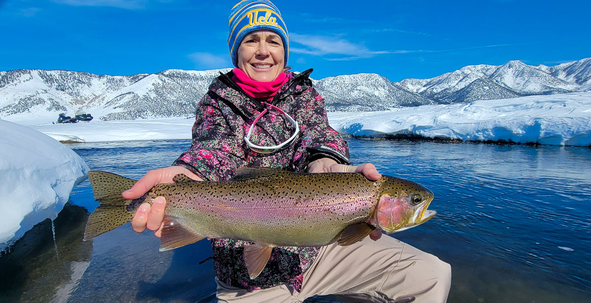 A smiling fisherwoman holding a massive rainbow trout next to a river in the snow in the eastern sierra fishing zone on the Upper Owens River.