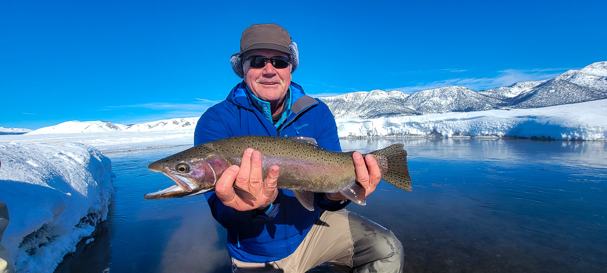 A smiling fisherman holding a massive rainbow trout next to a river in the snow in the eastern sierra fishing zone on the Upper Owens River.